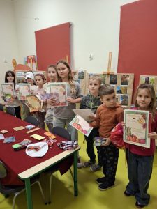 Grupa dzieci po wręczeniu nagród w szkolnym konkursie fotograficznym, trzymających dyplomy i upominki. W tle widoczna jest wystawa pokonkursowa z fotografiami przyrodniczymi umieszczonymi na tablicach.