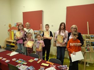 Grupa dzieci stojąca w sali szkolnej, trzymająca w rękach dyplomy za udział w konkursie fotograficznym. Obok nich stoją dwie dorosłe osoby – nauczycielka organizująca konkurs i pani dyrektor. W tle znajdują się plansze z wystawą pokonkursową przedstawiającą zdjęcia przyrodnicze.