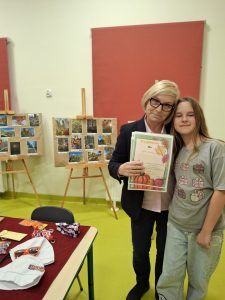 Dyrektor szkoły wręcza dyplom starszej uczennicy podczas podsumowania szkolnego konkursu fotograficznego. W tle widoczne są trzy tablice z wystawą nagrodzonych i wyróżnionych zdjęć.