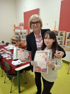 Dyrektor szkoły pozująca do zdjęcia z uczennicą trzymającą dyplom za wyróżnienie w konkursie fotograficznym. Obok stoi stół z nagrodami, a w tle znajduje się wystawa pokonkursowa pokazująca fotografie przyrodnicze wykonane przez uczniów.