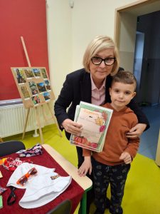 Dyrektor szkoły stojąca obok młodego ucznia, który trzyma dyplom za udział w szkolnym konkursie fotograficznym. Oboje pozują do zdjęcia w sali, w tle widać planszę z fotografiami pokonkursowymi oraz stół z nagrodami.