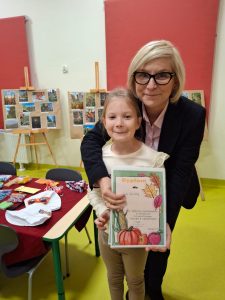 Dyrektor szkoły obejmuje uczennicę, która trzyma dyplom za wyróżnienie w szkolnym konkursie fotograficznym. Za nimi stoją tablice z prezentacją konkursowych fotografii oraz stół z nagrodami.