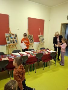 Dyrektor szkoły wręcza dyplom uczennicy, podczas gdy inni uczniowie stoją przy stole z nagrodami. W tle widoczne są plansze z fotografiami pokonkursowymi ustawione na sztalugach.