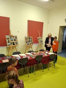 Dyrektor szkoły wręcza dyplom chłopcu w sali, w której ustawiono wystawę pokonkursową oraz stół z nagrodami i upominkami dla uczestników konkursu fotograficznego.