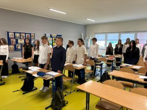 Cała klasa stoi przy ławkach podczas odśpiewania hymnu narodowego z okazji Święta Niepodległości. Uczniowie są ubrani odświętnie, a w tle widoczne są tablice i plakaty edukacyjne