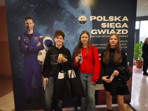 Troje uczniów stoi na tle wystawy z grafiką astronauty i napisem ‘Polska sięga gwiazd’. Każdy z nich trzyma pamiątkowe materiały z wydarzenia.