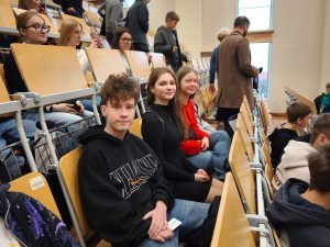 Uczniowie siedzą w pierwszych rzędach sali wykładowej, patrząc w stronę aparatu. Za nimi widoczni są inni uczestnicy spotkania zajmujący miejsca na trybunach