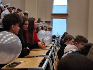 Uczniowie siedzą w dużej sali wykładowej, trzymając w rękach przezroczyste balony. W ławkach i na balkonach widać wielu młodych uczestników wydarzenia, skupionych na trwającym spotkaniu.