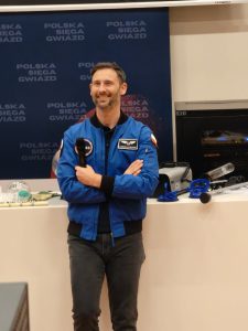 Prelegent w niebieskiej kurtce z naszywkami stoi przed stołem demonstracyjnym na tle planszy ‘Polska sięga gwiazd’. Uśmiecha się i trzyma mikrofon.