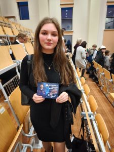 Uczennica stoi w sali wykładowej i trzyma pamiątkową kartę z grafiką wydarzenia ‘Polska sięga gwiazd’. W tle znajdują się krzesła oraz inni uczestnicy opuszczający aulę.