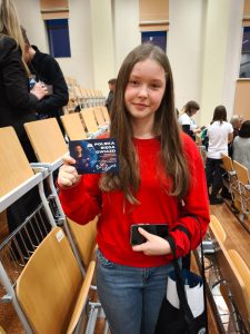 Uczennica w czerwonym swetrze stoi w auli i prezentuje kartę pamiątkową otrzymaną podczas wydarzenia ‘Polska sięga gwiazd’. W tle widać krzesła oraz wychodzących uczestników.