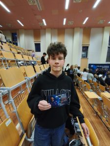 Uczeń stoi w sali wykładowej i trzyma w ręku pamiątkową kartę z grafiką przedstawiającą uczestnika wydarzenia ‘Polska sięga gwiazd’. Za nim widoczne są rzędy pustych oraz zajmowanych krzeseł.