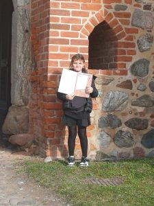Uczennica Szkoły Podstawowej w Ostaszewie stoi przed ceglano-kamiennym murem i trzyma w dłoniach otwartą teczkę z dyplomem. Dziewczynka pozuje do zdjęcia na tle architektury z czerwonej cegły. Fotografia wykonana w związku z udziałem w wojewódzkim konkursie plastycznym ‘Czas zostawił ślad…’