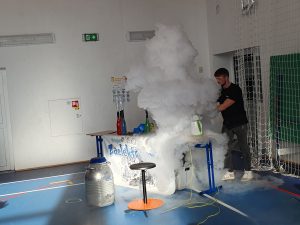 Pokaz doświadczenia z suchym lodem – prowadzący stoi przy stole, z którego wydobywa się duża chmura białej pary, a uczniowie obserwują eksperyment.