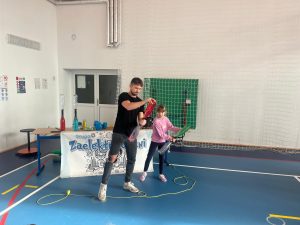 Prowadzący pokaz naukowy pomaga uczennicy przeprowadzić doświadczenie z butelkami wypełnionymi kolorową cieczą. Oboje trzymają butelki i obserwują działanie eksperymentu na sali gimnastycznej.