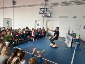 Prowadzący siedzi na obrotowym stołku i trzyma w dłoni płonący przedmiot podczas doświadczenia z ogniem, a grupa dzieci siedzi przed nim na ławkach i z zainteresowaniem obserwuje pokaz.