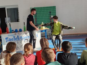 Uczestnik pokazu siedzi na obrotowym stołku z rozłożonymi rękami, trzymając hantle, a prowadzący tłumaczy zasadę działania doświadczenia z momentem obrotowym.