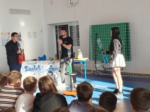 Pokaz naukowy w sali gimnastycznej – prowadzący oraz dwoje uczniów trzymają butelki z kolorową cieczą, a przed nimi siedzi grupa uczniów oglądających doświadczenie.