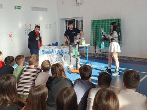 Pokaz naukowy – dwoje uczniów trzyma butelki z kolorową cieczą, a prowadzący objaśnia doświadczenie. Uczniowie siedzą na sali gimnastycznej i uważnie oglądają eksperyment.