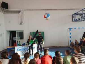 Podczas pokazu naukowego uczennica trzyma urządzenie wyrzucające powietrze, a w powietrzu unosi się kolorowa piłka plażowa. Obok stoi prowadzący, a uczniowie siedzą i z zainteresowaniem obserwują eksperyment.