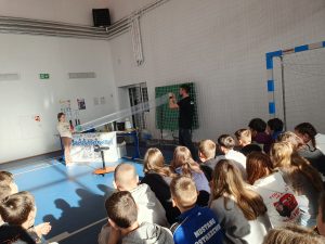 Prowadzący pokaz naukowy wraz z uczennicą stojąca na przeciwko trzymają napełniony powietrzem długi worek foliowy. Pozostali uczniowie siedzą na sali i obserwują doświadczenie.