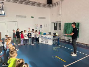 Grupa dzieci stoi w szeregu przed prowadzącym, który pokazuje doświadczenie z długi workiem foliowym wypełnionym powietrzem. Pozostałe dzieci siedzą na podłodze i obserwują pokaz.