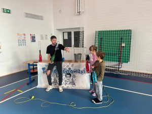 Prowadzący trzyma butelkę z zieloną cieczą i tłumaczy doświadczenie dwójce dzieci stojących obok niego, którzy trzymają podobne butelki z kolorową wodą.