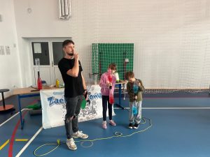 Prowadzący pokaz naukowy stoi obok dwojga dzieci, które trzymają butelki z kolorową cieczą i wykonują doświadczenie. Za nimi znajduje się stół z przyrządami i plakat grupy „Zaelektryzowani”.