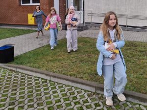Kilka uczennic idzie po trawniku w kierunku aparatu, niosąc w rękach karmę dla zwierząt. W tle widać budynek szkoły i osobę dorosłą wychodzącą na chodnik.