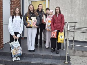 Grupa uczniów stoi na schodach przed budynkiem szkolnym, trzymając torby i worki z karmą dla zwierząt. Wszyscy mają zimowe ubrania, a w rękach różne rodzaje karmy dla psów i kotów