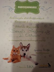 Dyplom z podziękowaniem od Schroniska dla Bezdomnych Zwierząt w Toruniu dla Szkoły Podstawowej za darowiznę karmy. Na dole ilustracja kota i psa oraz podpis kierownika schroniska.