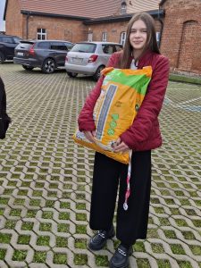 Uczennica stojąca na parkingu przed budynkiem z czerwonej cegły, trzymająca w obu rękach duży worek karmy dla psa. W tle widoczne są zaparkowane samochody.