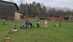 Grupa dzieci bawi się na świeżym powietrzu na terenie placówki, pchając zabawkowy pojazd po trawie.