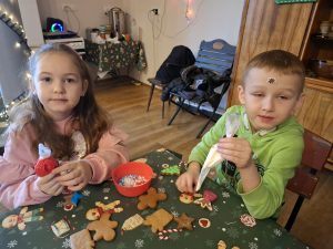 Dwoje dzieci trzyma rękawy cukiernicze i ozdabia pierniki przy świątecznie nakrytym stole w sali przedszkolnej.