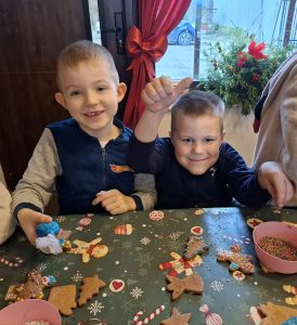 Dwoje dzieci z oddziału przedszkolnego siedzi przy stole i dekoruje pierniki lukrem oraz kolorowymi posypkami podczas warsztatów świątecznych.