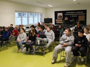 Uczniowie klas 4 i 5 siedzą na krzesłach, uważnie słuchając wystąpienia gościa w szkolnej sali.