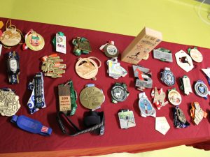 Zbliżenie na stół przykryty czerwonym obrusem, na którym rozłożono liczne medale, identyfikatory i pamiątki z maratonów.