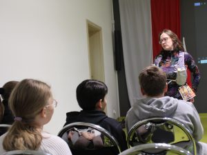 Prelegentka w stroju biegowym rozmawia z kilkoma uczniami, którzy siedzą w pierwszym rzędzie.
