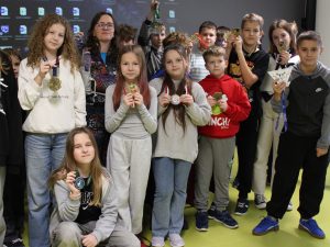 Grupa uczniów pozuje wspólnie z prelegentką, trzymając w dłoniach różne medale biegowe zdobyte podczas spotkania.