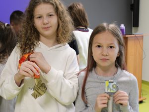 Dwie dziewczynki pozują z medalami, trzymając je przed sobą i uśmiechając się do aparatu.