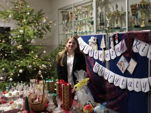 Młoda osoba stoi za stoiskiem kiermaszu bożonarodzeniowego z rękodziełem i zapakowanymi prezentami. W tle widoczna choinka i baner kiermaszu.