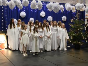 Grupa dziewcząt ubranych w białe stroje aniołów stoi na scenie. W tle świąteczne choinki oraz białe papierowe ozdoby zawieszone nad sceną.
