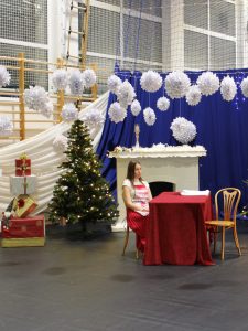 Dziewczynka w świątecznym stroju siedzi przy czerwonym stole na tle dekoracyjnego kominka i choinki. Nad sceną wiszą białe papierowe ozdoby.