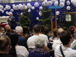 Wójt gminy Łysomice przemawia do licznie zgromadzonej publiczności. Za nim znajduje się scenografia szkolnych Jasełek: choinki, światełka i dekoracja stylizowana na pociąg.