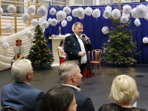 Przewodniczący Rady Gminy Łysomice Robert Kożuchowski stojący na scenie z mikrofonem przemawia do publiczności. W tle dekoracje świąteczne: choinki, białe papierowe ozdoby i niebieska kurtyna z lampkami.