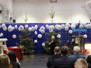 Pani dyrektor stojąca na scenie czyta z kartki. Za nią świąteczne dekoracje, duże choinki z lampkami i zawieszone białe ozdoby. Publiczność siedzi przed sceną.