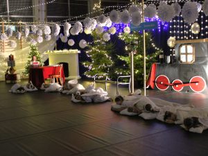 Grupa dzieci przebranych za aniołki leży na scenie na białych poduszkach. Obok znajduje się dekoracyjny kominek, choinki oraz biało-niebieskie oświetlenie.