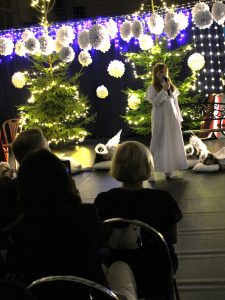 Dziewczynka w białej szacie stoi na scenie z mikrofonem. Za nią leżą dzieci w strojach aniołów oraz stoją świąteczne choinki.