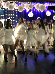 Grupa dziewcząt w białych strojach wykonuje taniec na scenie. W tle wiszą białe ozdoby oraz światełka, tworząc świąteczną dekorację.