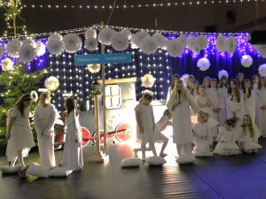 Grupa dzieci w białych strojach aniołów stoi na scenie. Jedno z dzieci trzyma mikrofon i przemawia. W tle widoczne są światełka oraz element dekoracyjny stylizowany na pociąg.